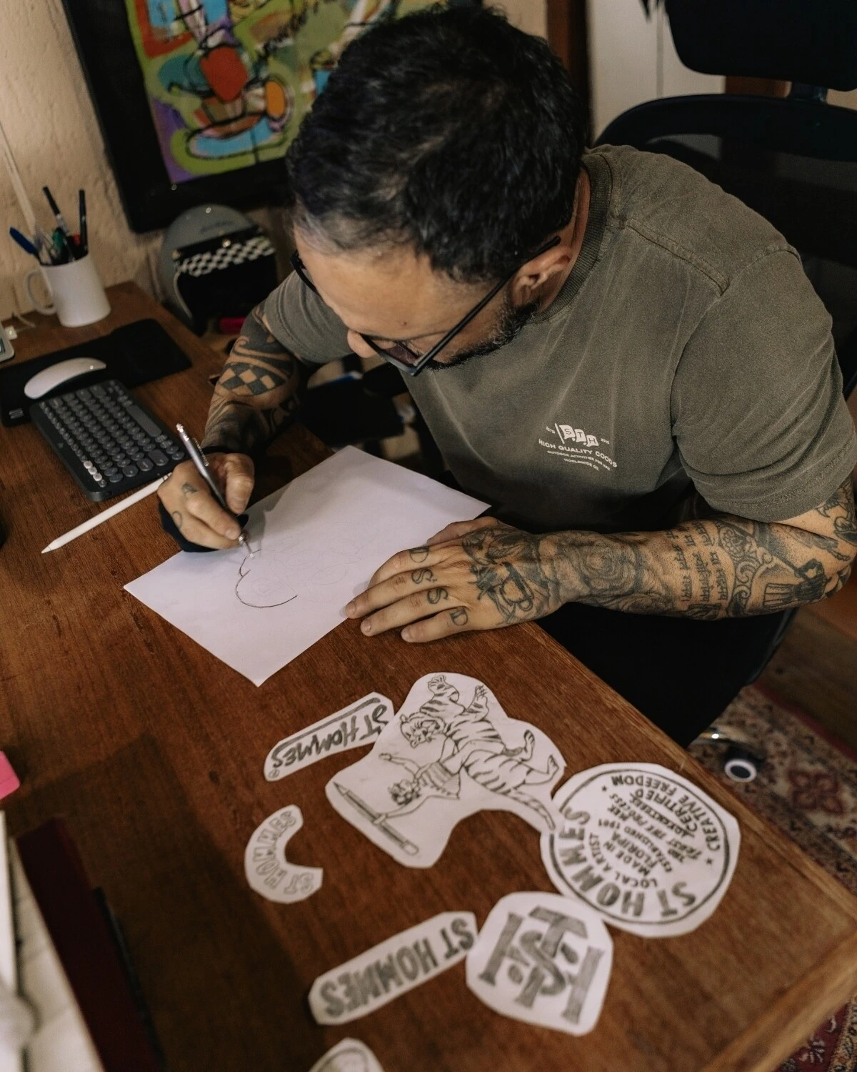 Fabio Luiz desenhando a lápis em uma folha, com os desenhos recortados sobre a mesa