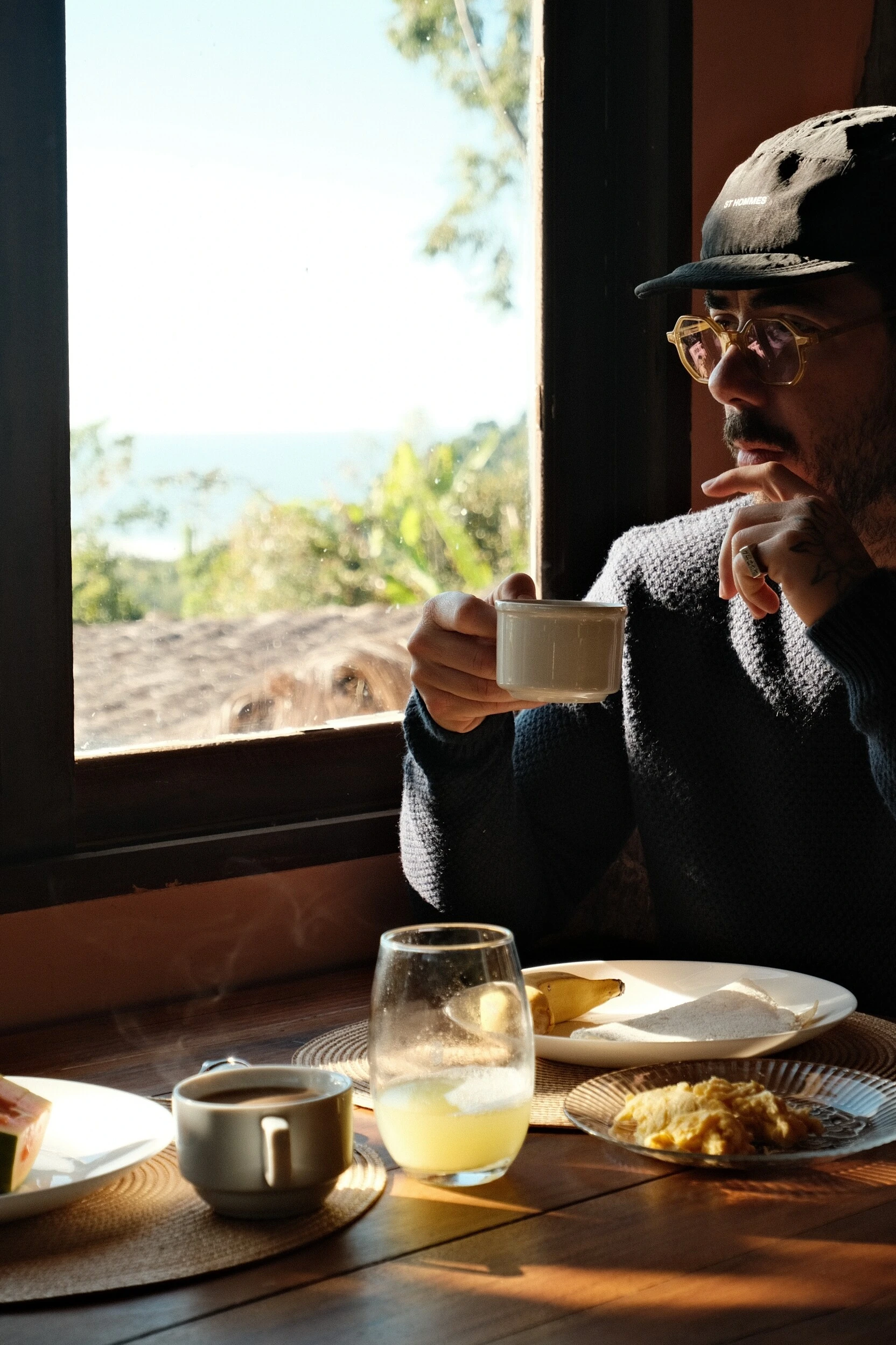 Murilo tomando café diante de uma janela com vista para a mata