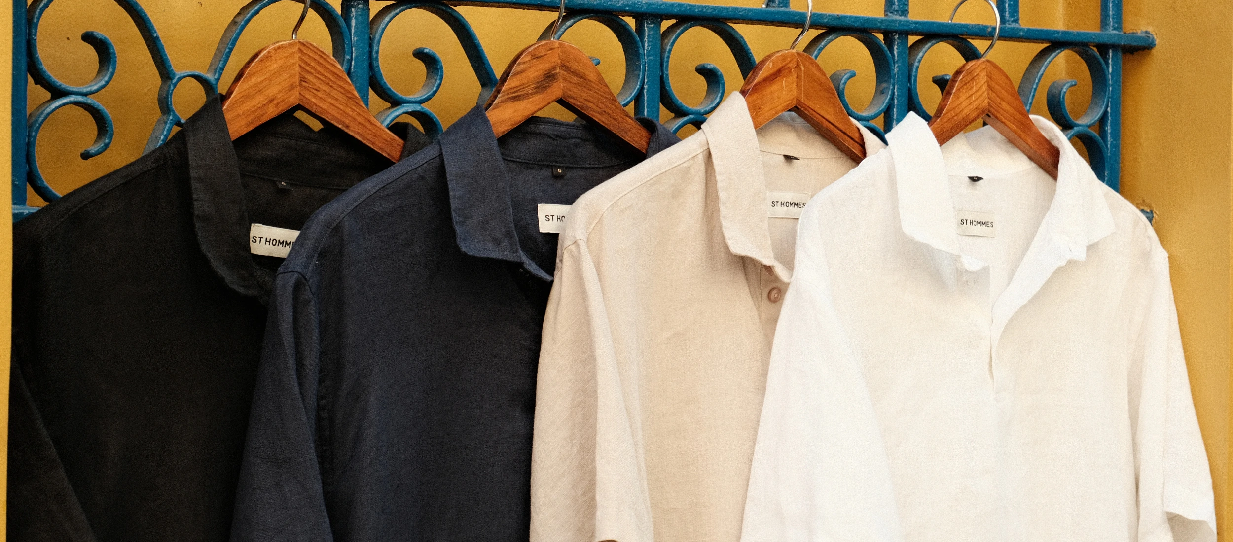 Quatro camisas de linho penduradas em portão de ferro ornamentado: preta, azul-marinho, areia e branca