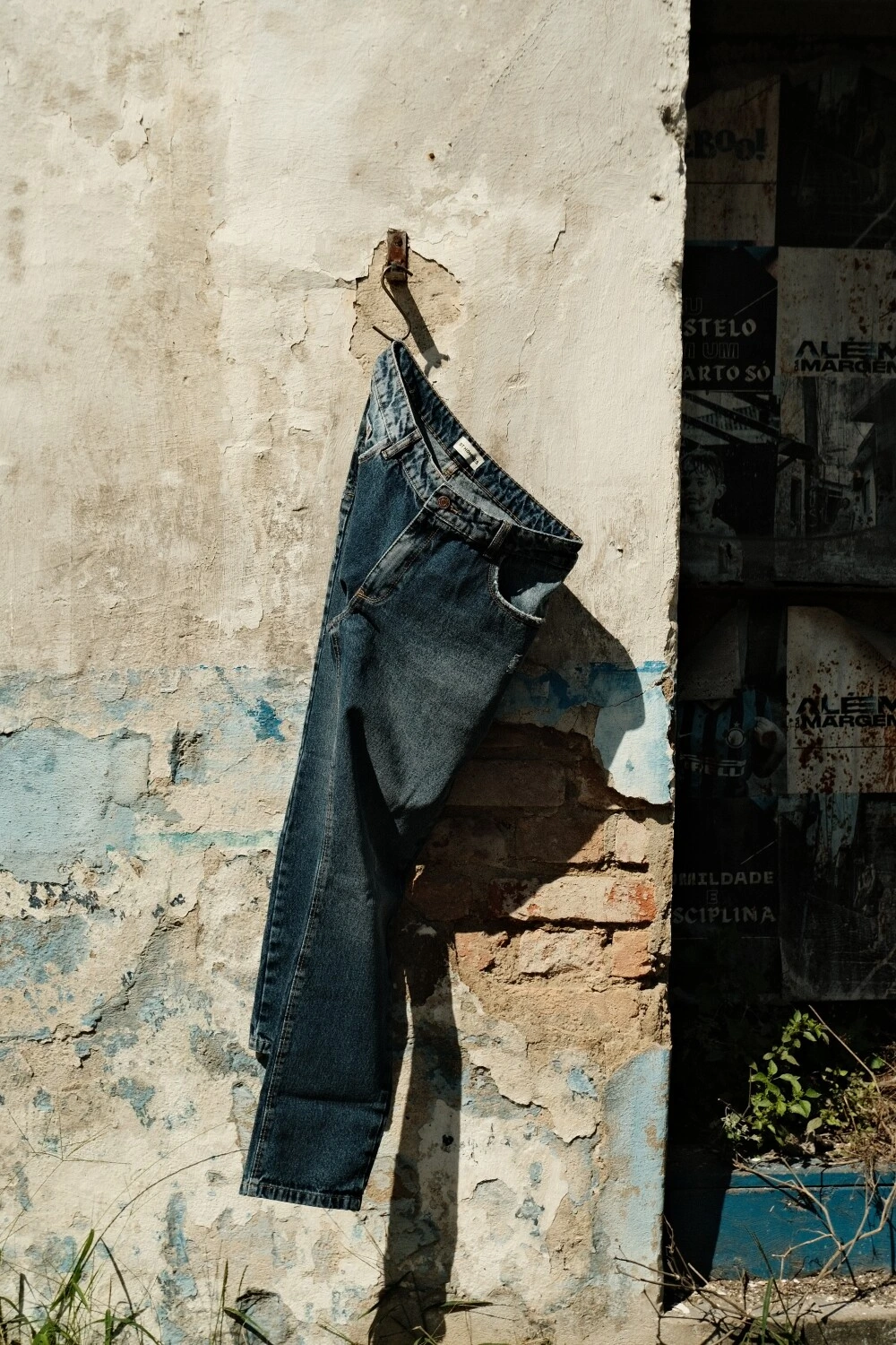 Calça jeans pendurada em gancho, contra parede descascada com tijolo aparente