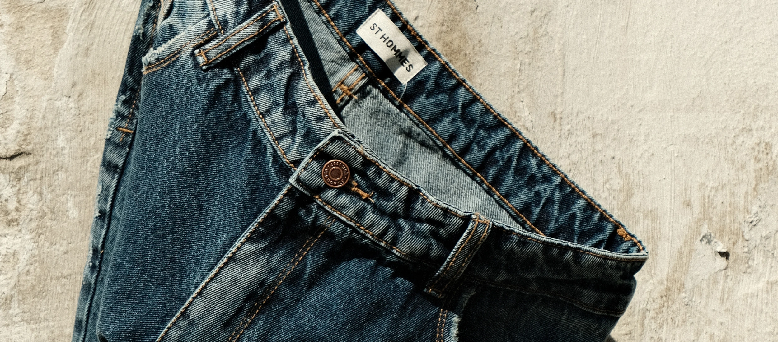 Detalhe do botão de metal e etiqueta ST HOMMES na cintura da calça jeans